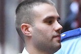 A man with short brown hair, in a white collared shirt, is escorted into a building by prison guards.