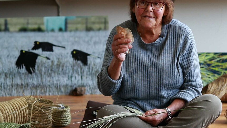 Woman sits on floor surrounded by woven baskets, holding kangaroo bone