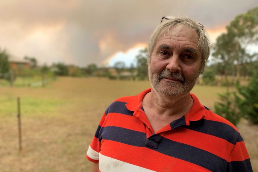 A man stands in a dry paddock in front of an orange and smoky sky.