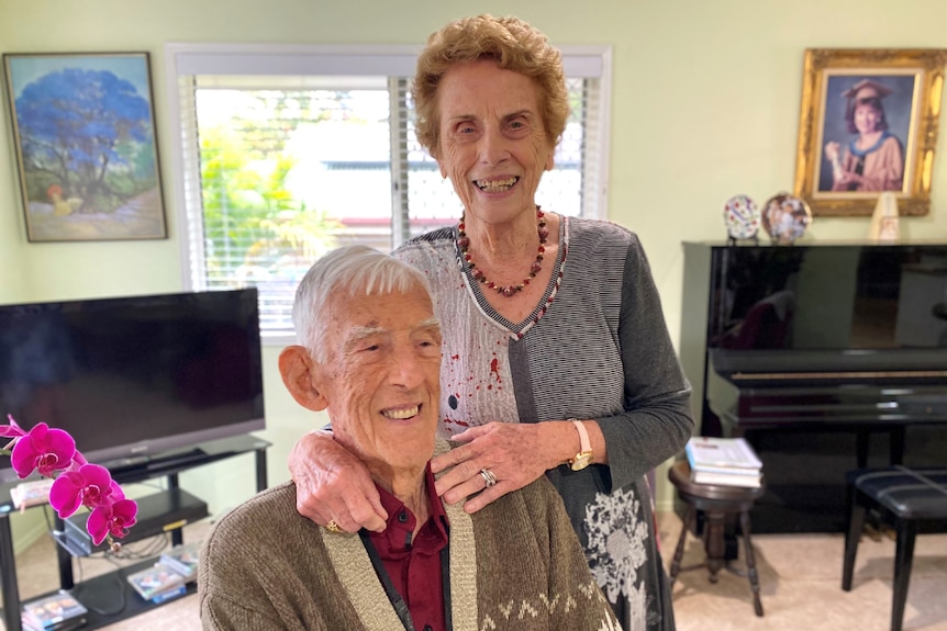 Woman in her 80 stands beside man who is 100, he's sitting and they're smiling