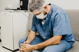 Man dressed in a nurse's scrubs wearing a hair net and face mask sitting down and looking down at his hands. 