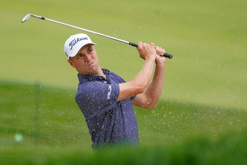 A golfer finishes his swing out of a bunker, with sand spraying in the air.