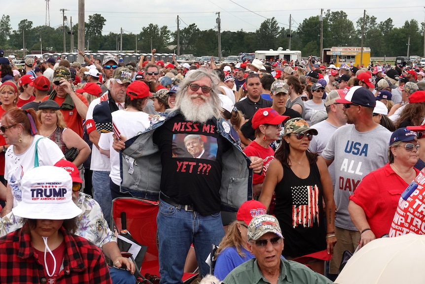 A crowd of people stands around, many with red caps. A man opens his vest to show a Trump t-shirt with the words 'miss me yet?'