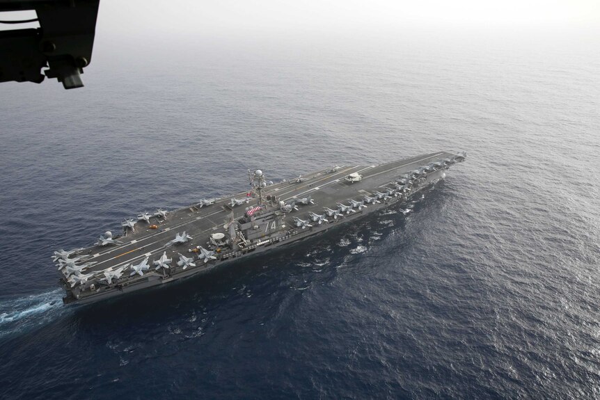 An aerial photo of an aircraft carrier with a host of fighter jets parked on board sails in the open sea