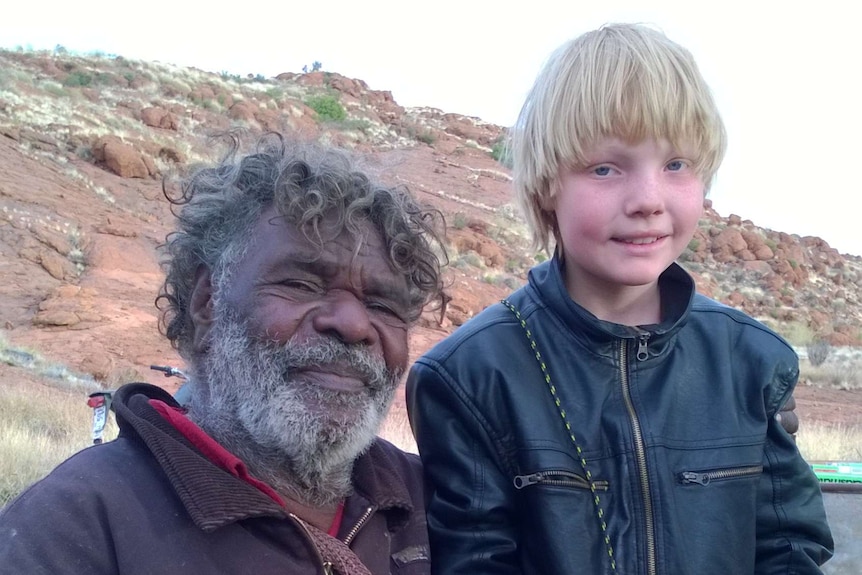 An older man smiles for a photo with a young blonde-haired boy in a black jacket in front of red rocks.