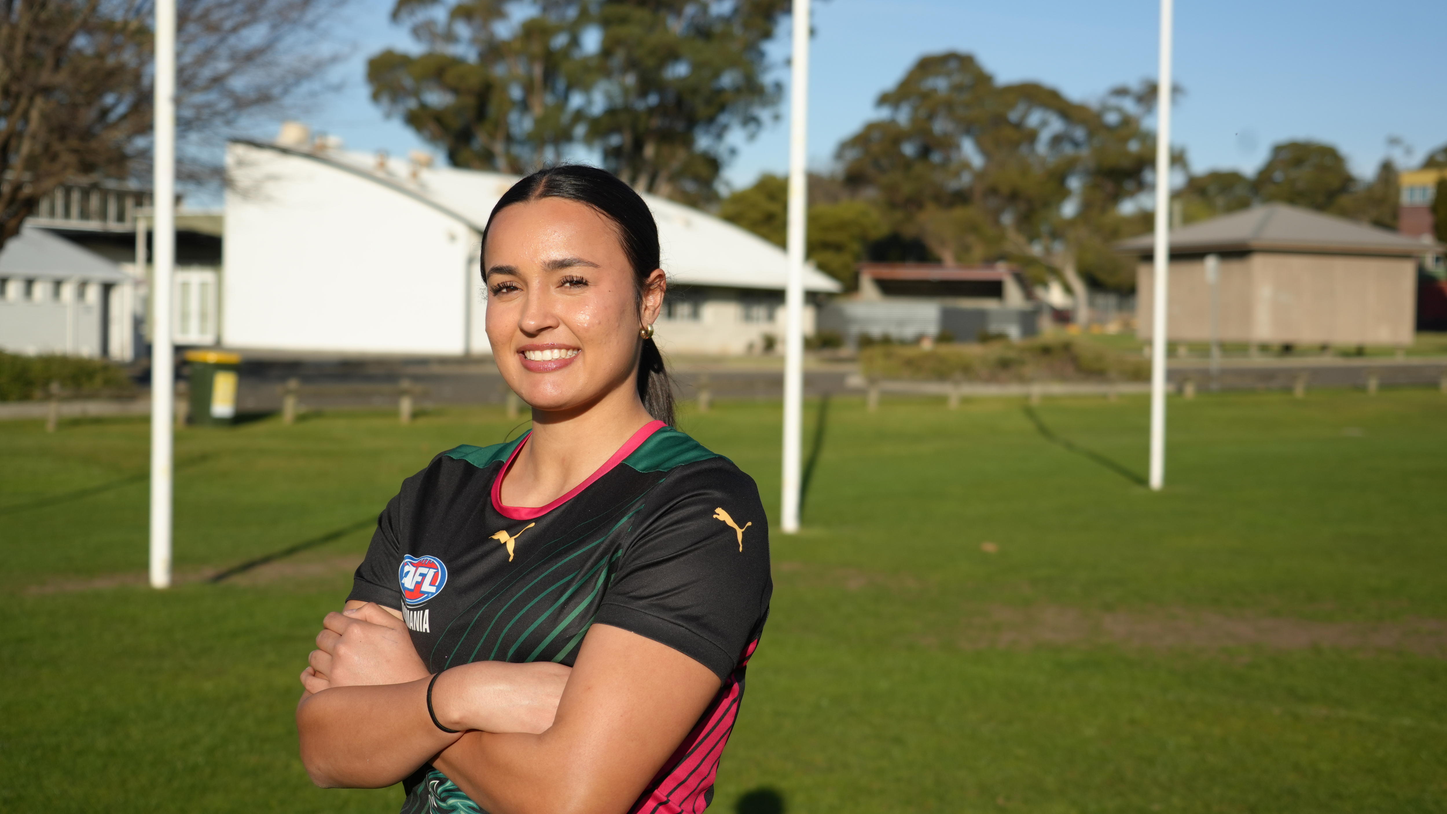 A young woman with dark hair standing with her arms crossed smiling in front of footy goals.