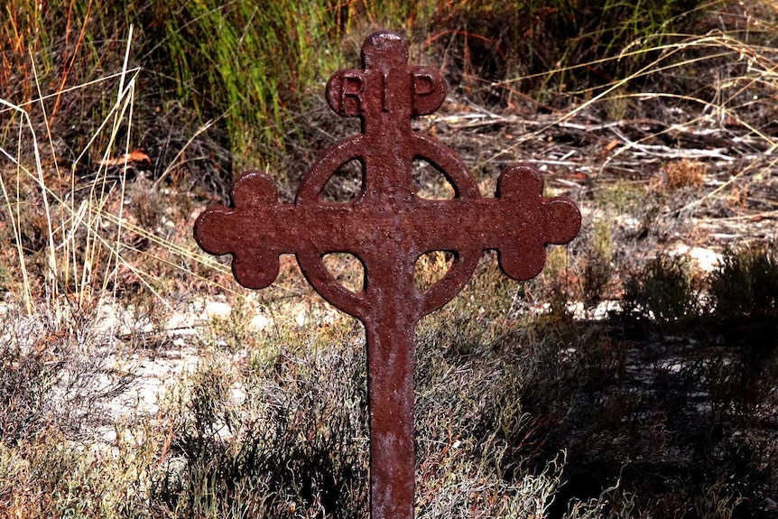 A rusty iron cross with the words RIP on the top with a scrubby grass backdrop.