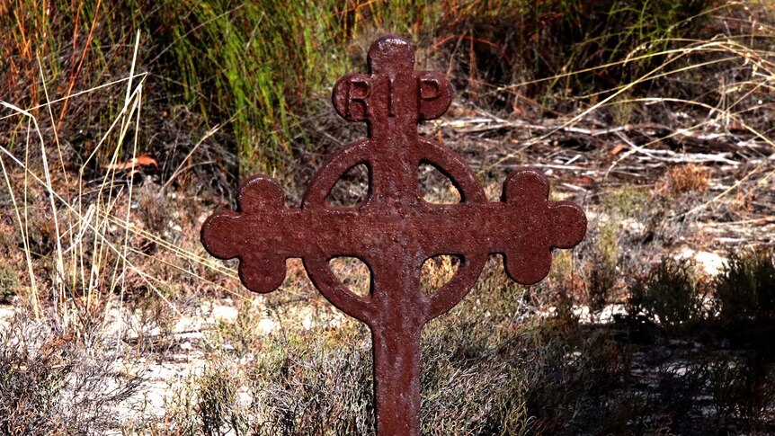 A rusty iron cross with the words RIP on the top with a scrubby grass backdrop.