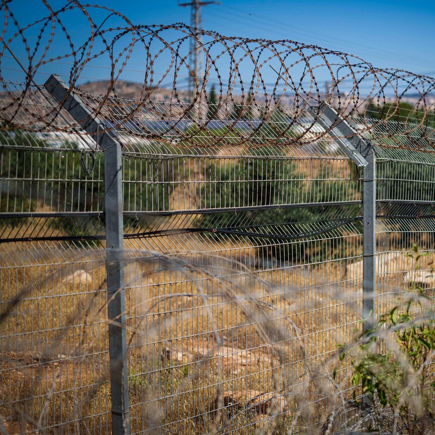 A barbed wire fence surrounds dry brown grass and green bushes, against a blue sky