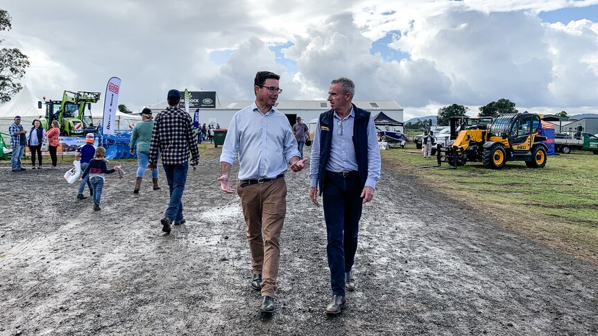 Two men walk side by side along a muddy road at a showground beneath an overcast sky.