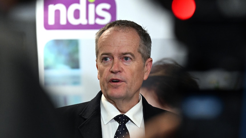 Bill Shorten stands in front of a sign displaying an NDIS logo. He is wearing a black suit and tie.