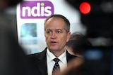 Bill Shorten stands in front of a sign displaying an NDIS logo. He is wearing a black suit and tie.