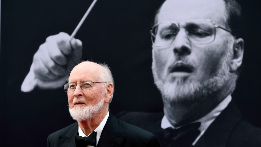 John Williams stands in front of a projected black and white image of himself.