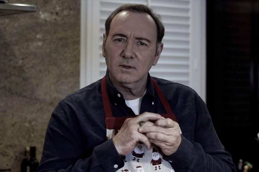 Kevin Spacey, wearing a Christmas-themed apron, puts a ring onto his finger while starting at the camera.