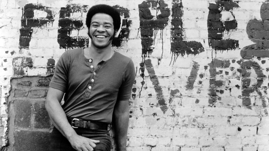 A black and white photo of a man in t-shirt and jeans standing in front of a graffiti wall. He has a big smile.