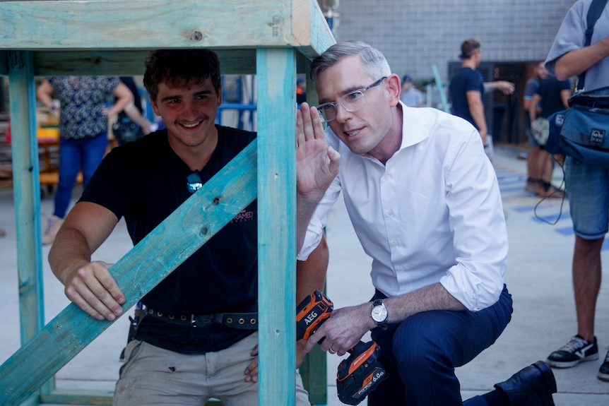 a man wearing glasses kneeling down next to a young man and working on a wooden table