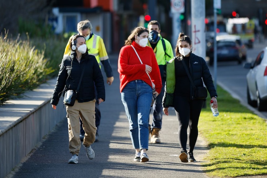 Tres mujeres jóvenes con máscaras caminando por una calle de Melbourne.  Detrás de ellos hay dos hombres con ropas altas y máscaras.