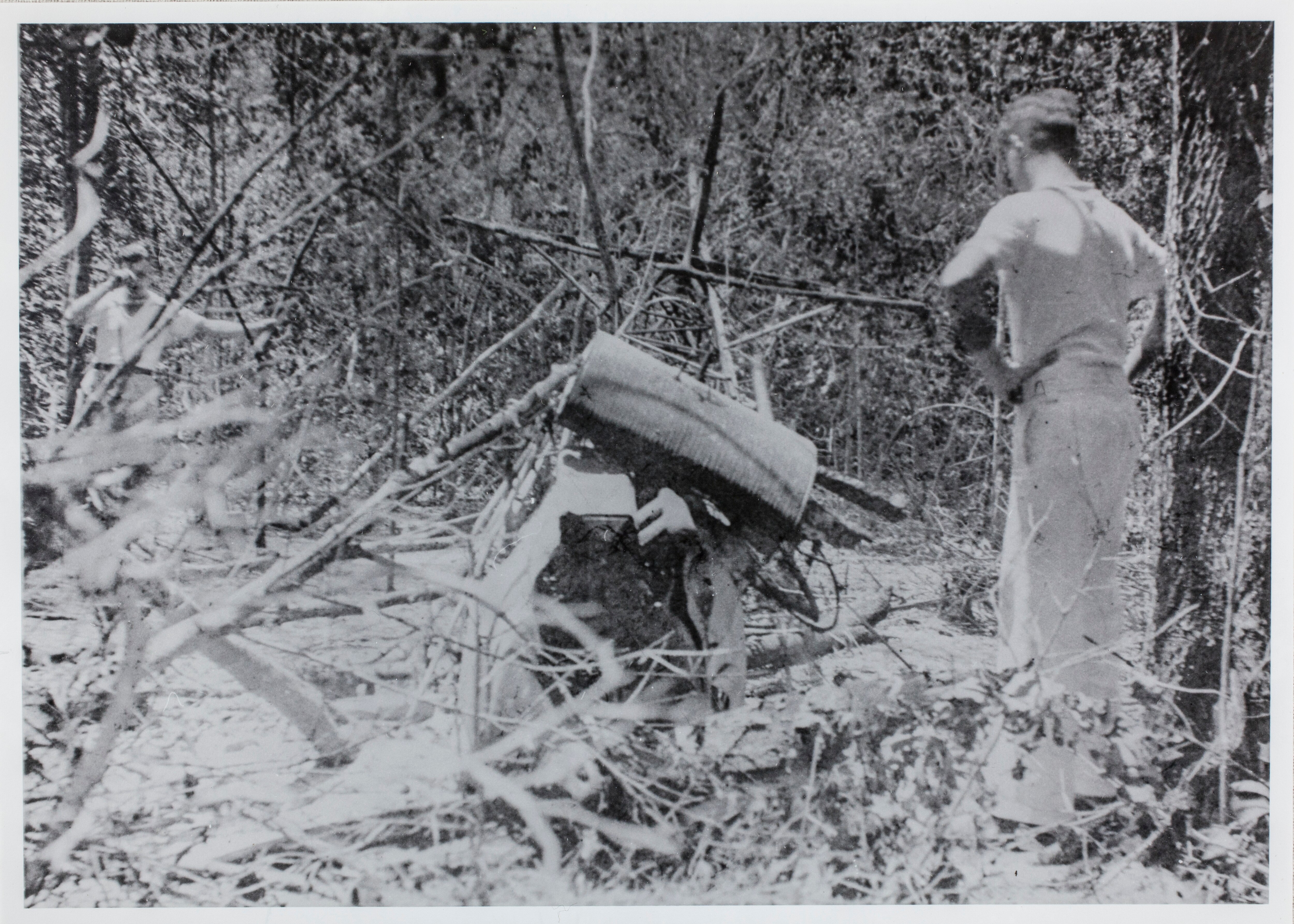 A man looks at an old airplane on the ground in the bush.  The photo is old and in black and white. 