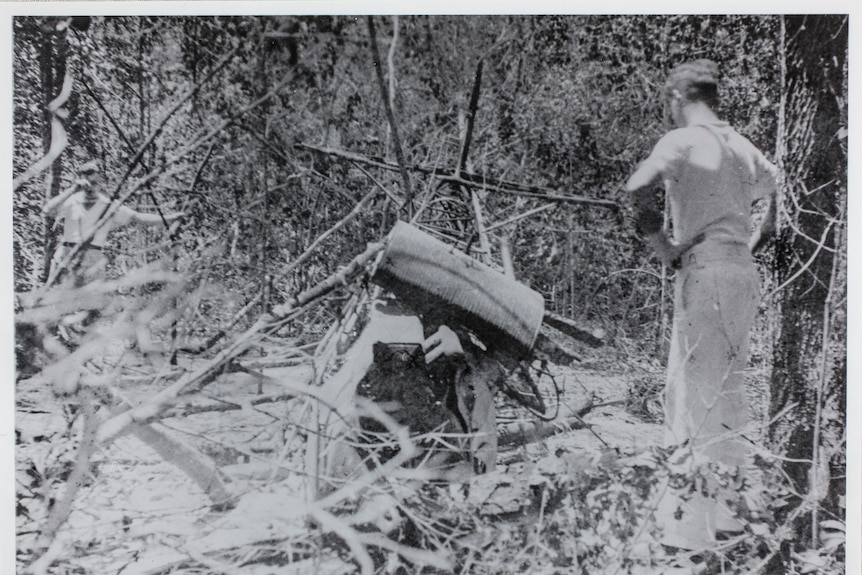 A man looks at an old airplane on the ground in the bush.  The photo is old and in black and white. 