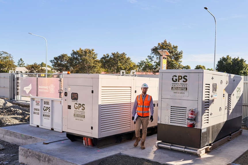 A man in an orange safety vest walks between two large generators. 