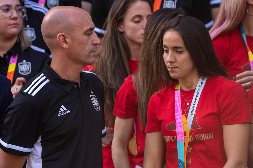 A man in a black polo stands, looking at a woman in a red shirt with a medal around her neck