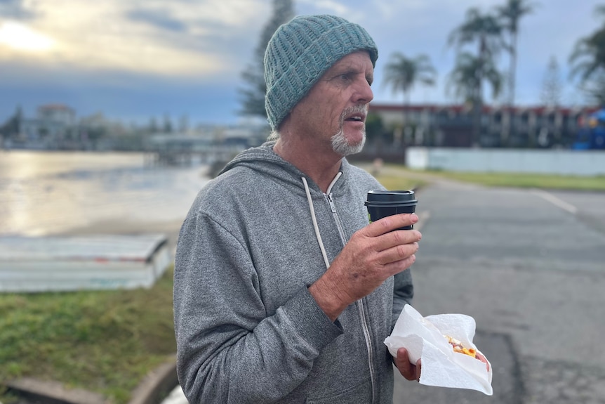 A man wearing a warm top and beanie holds a cup of coffee, with a river in the background.