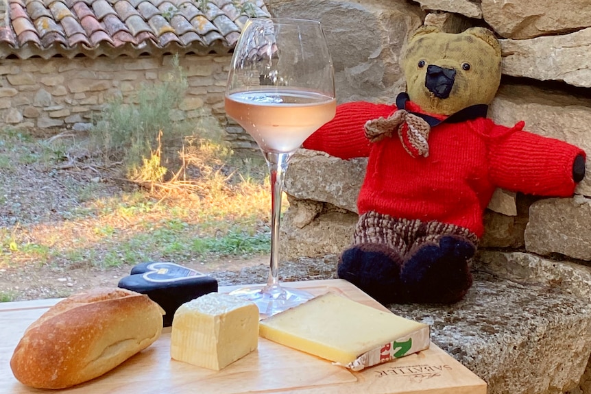 A teddy bear with a red jacket sits next to a platter of cheese, a bread roll and wine.