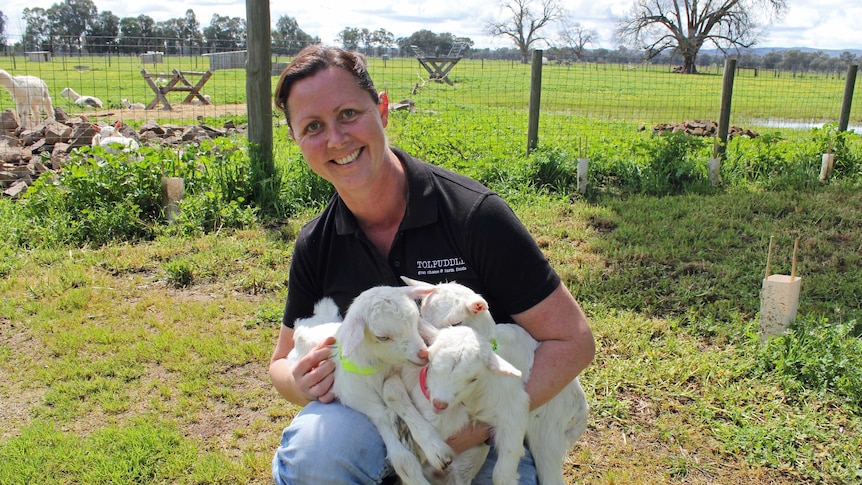 Melissa kneels down with three kid goats in her arms with the farm behind her