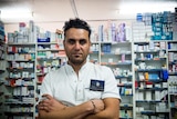 A pharmacist in a white coat standing in front of shelves of medications in a pharmacy.