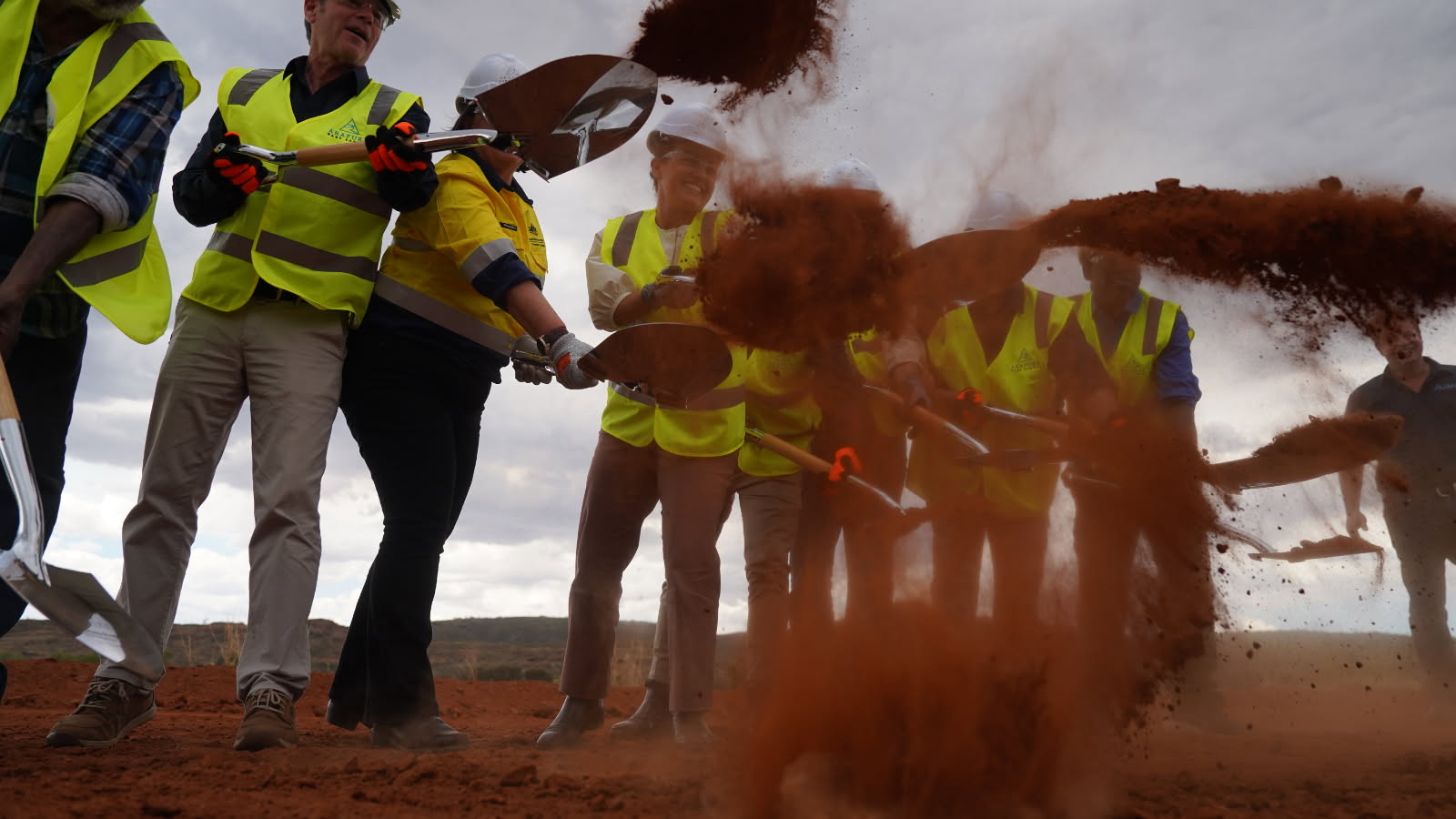 Red dirt flies in the air from shovels, with people seen through the dirt wearing high vis vests