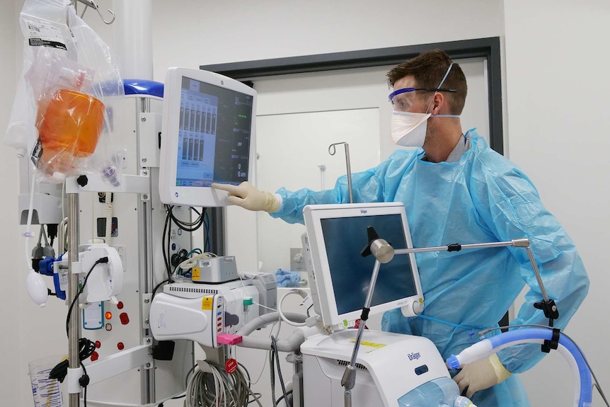 A nurse wearing a mask and gown, touches a screen with medical information