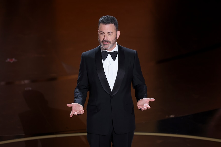 Jimmy Kimmel with hands out, wearing a suit and bow tie, standing on stage, brown background