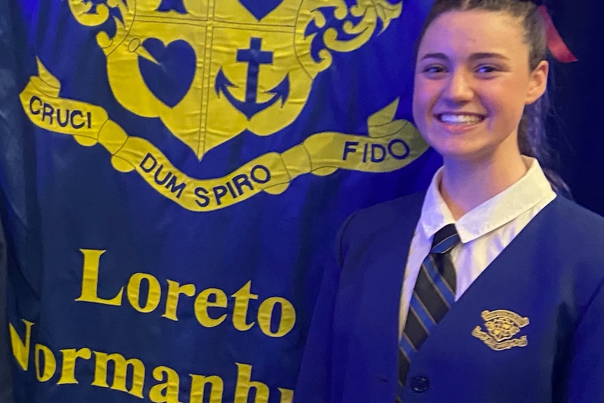 A girl in a blue and yellow school uniform smiles at the camera next to a school flag with crest.
