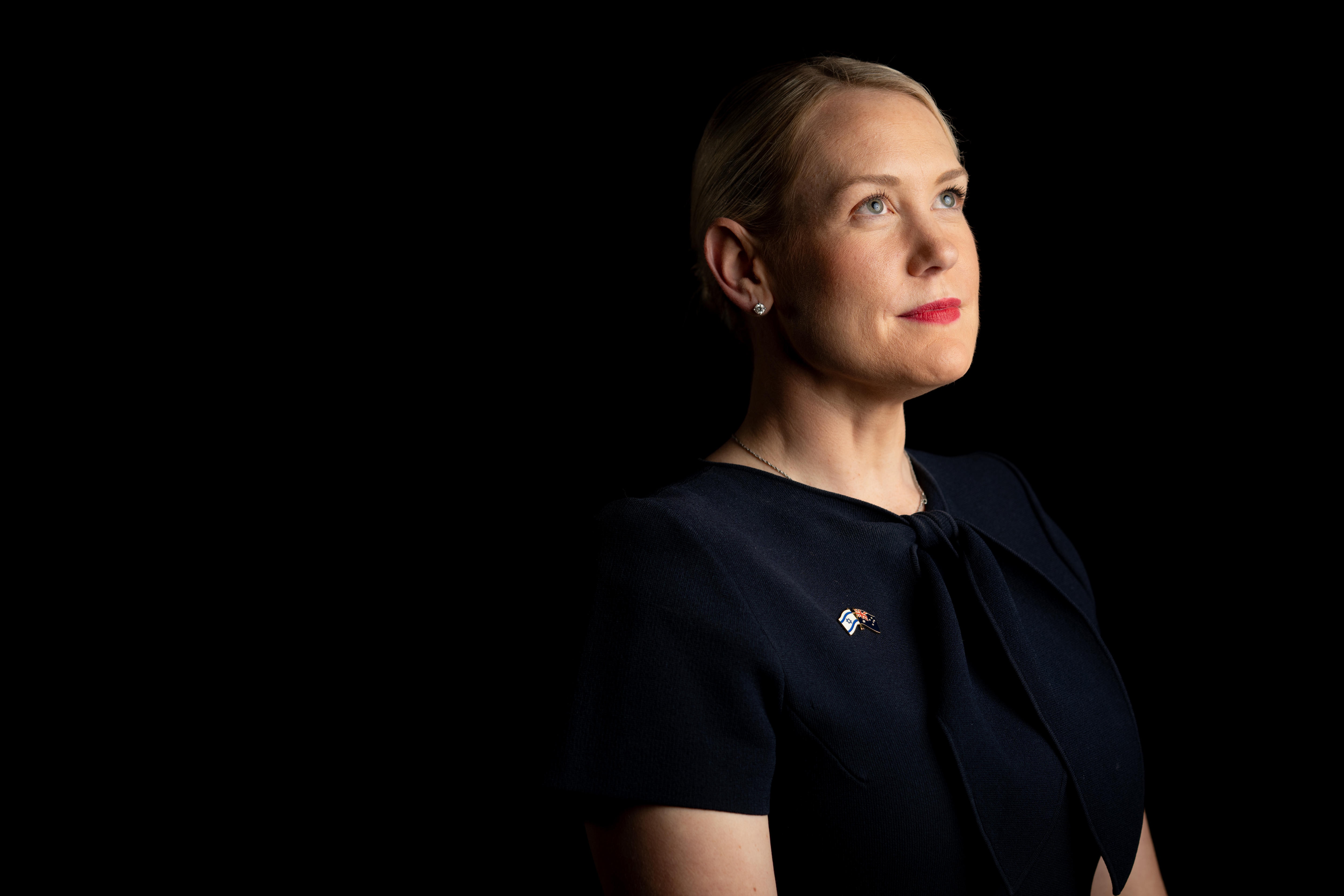 Uma mulher de cabelos loiros e vestido azul marinho olha para cima com um leve sorriso contra um fundo escuro.