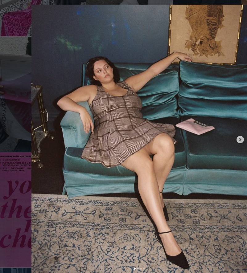  A plus-size woman reclines on a turquoise velvet sofa, looks away, wears brown check short dress, auburn hair.