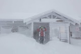 A skier waiting in a doorway as snow falls on Mt Hotham, and snow piles up against a wooden house.