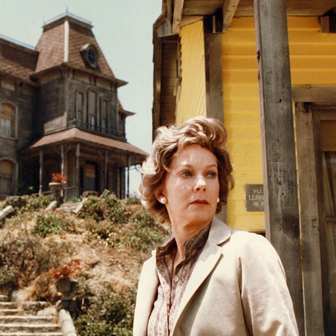 A woman with auburn hair in a short, wavy style with a serious expression standing in front of a scary-looking old house.