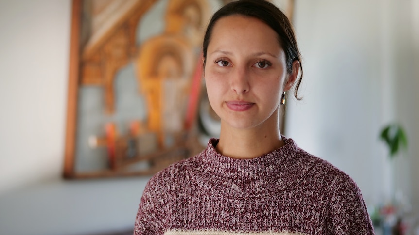 A young woman with dark hair stands in her house, looking solemn.