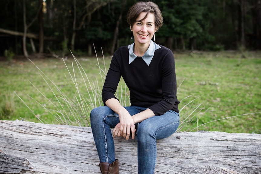A woman with short brown hair smiles while sitting on a log.