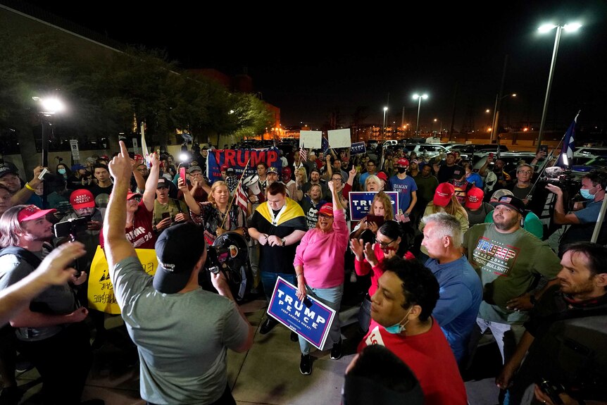 A man with a reverse baseball cap and megaphone leads a chant of sign-waving protesters at night.