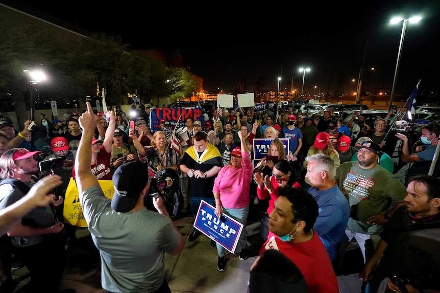 A man with a reverse baseball cap and megaphone leads a chant of sign-waving protesters at night.