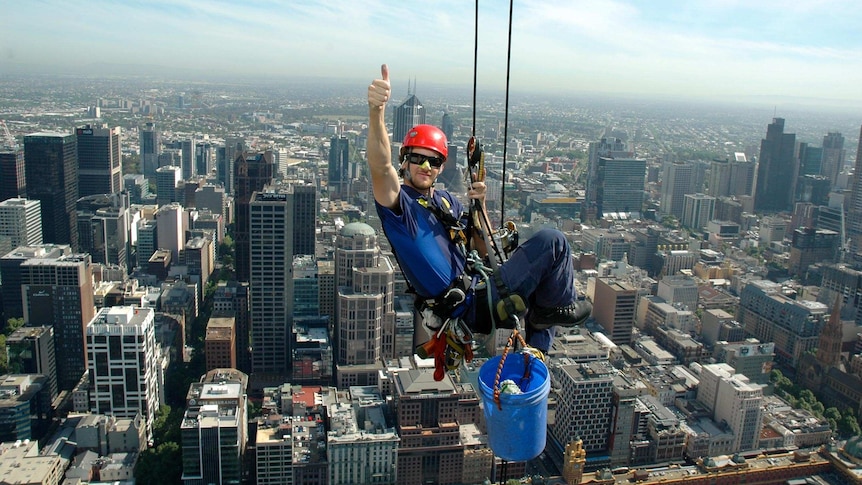 A man abseils from a building with a vista of Melbourne behind him.