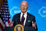 Joe Biden speaks behind a lectern with the US Presidential seal, in front of a blue digital screen.