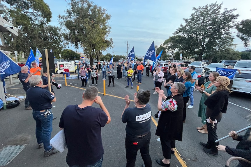 About 30 protestors stand in a carpark in a rough circle. Some are holding large blue flags Australian Services Union flags.