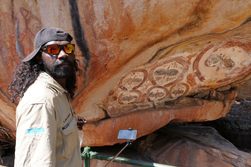 indigenous man with selfie stick stands in front of rock art