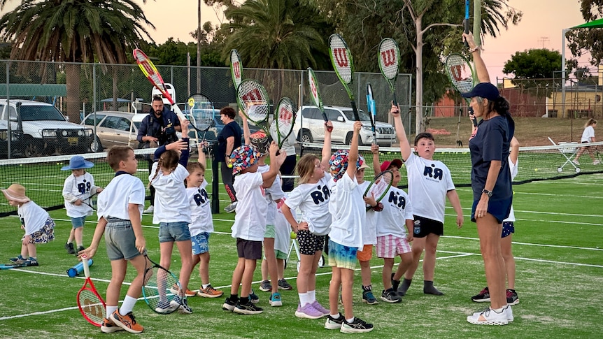 A group of children hold tennis racquets above their heads as they watch their tennis coach do the same