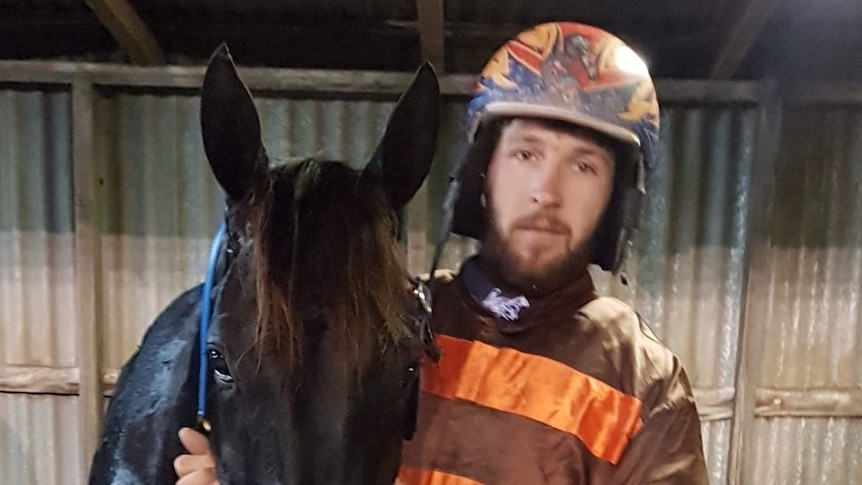 A bearded man wearing a crash helmet and racing gear posing with a horse.