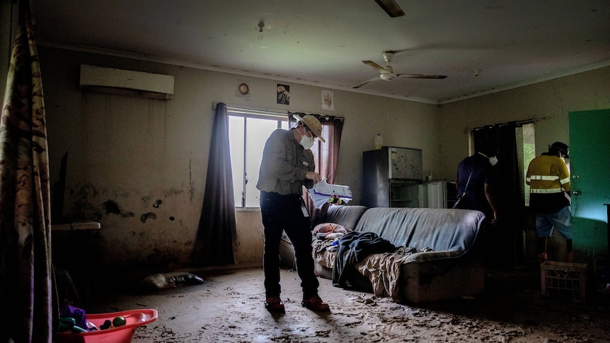 A man in a facemask with a clipboard stands in a muddy lounge room
