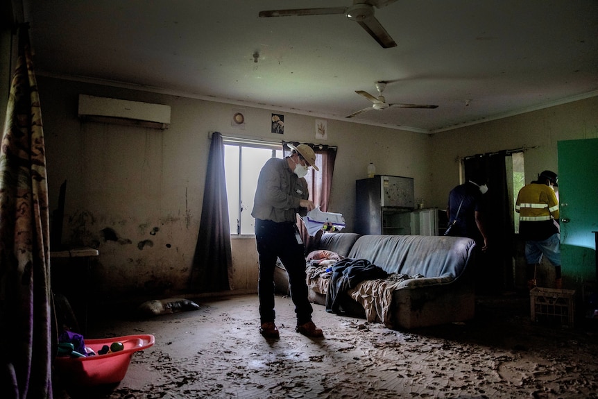 A man in a facemask with a clipboard stands in a muddy lounge room