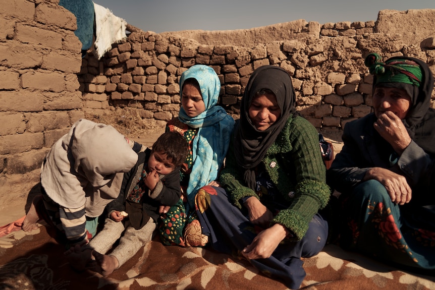 Two women and several children sit in a mudbrick courtyard.